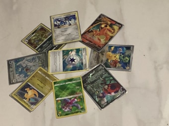A collection of Pok&eacute;mon trading cards is arranged in a circular pattern on a white surface. The cards feature various Pok&eacute;mon characters, each with different illustrations and designs. Some cards have a metallic or holographic finish, adding a reflective quality to the images.