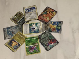 A collection of Pokémon trading cards is arranged in a circular pattern on a white surface. The cards feature various Pokémon characters, each with different illustrations and designs. Some cards have a metallic or holographic finish, adding a reflective quality to the images.