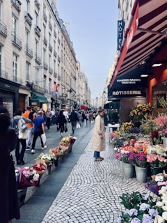 A bustling, narrow street lined with historic buildings and vibrant shops. People stroll along the cobblestone path, some carrying shopping bags. A flower stall with a variety of colorful bouquets is prominently displayed in the foreground, adding charm and liveliness to the scene. The awnings and signage on the buildings suggest a mix of retail and dining establishments. The sky is clear, indicating pleasant weather.