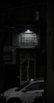 A dimly lit street scene features a parked car partially visible under a streetlight, casting shadows on the building facade. The building has a grid-like window and a door with advertisement posters. The atmosphere is dark, suggesting nighttime.