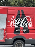 A red delivery truck with a large Coca-Cola logo and the phrase 'Real Magic' printed on the side. The design includes two black silhouette bottles next to the brand name. The truck's door is partially open, revealing a metallic interior. The background features green foliage.