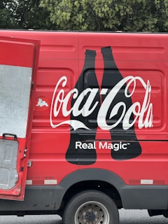 A red delivery truck with a large Coca-Cola logo and the phrase 'Real Magic' printed on the side. The design includes two black silhouette bottles next to the brand name. The truck's door is partially open, revealing a metallic interior. The background features green foliage.