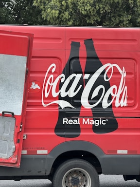 A red delivery truck with a large Coca-Cola logo and the phrase 'Real Magic' printed on the side. The design includes two black silhouette bottles next to the brand name. The truck's door is partially open, revealing a metallic interior. The background features green foliage.