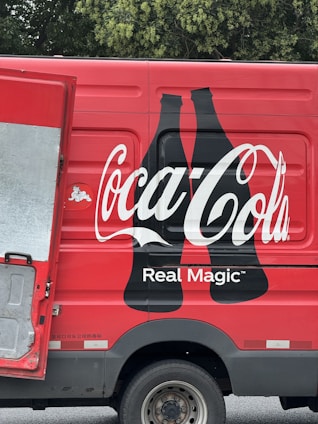 A red delivery truck with a large Coca-Cola logo and the phrase 'Real Magic' printed on the side. The design includes two black silhouette bottles next to the brand name. The truck's door is partially open, revealing a metallic interior. The background features green foliage.