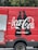 A red delivery truck with a large Coca-Cola logo and the phrase 'Real Magic' printed on the side. The design includes two black silhouette bottles next to the brand name. The truck's door is partially open, revealing a metallic interior. The background features green foliage.