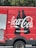 A red delivery truck with a large Coca-Cola logo and the phrase 'Real Magic' printed on the side. The design includes two black silhouette bottles next to the brand name. The truck's door is partially open, revealing a metallic interior. The background features green foliage.