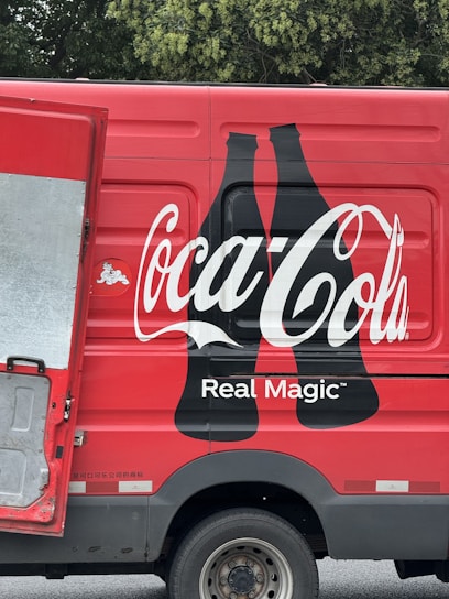 A red delivery truck with a large Coca-Cola logo and the phrase 'Real Magic' printed on the side. The design includes two black silhouette bottles next to the brand name. The truck's door is partially open, revealing a metallic interior. The background features green foliage.