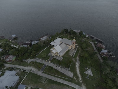 An aerial view of a church with a large roof and a prominent bell tower topped with a cross, surrounded by lush green vegetation and a few other structures. The complex is adjacent to a large body of water, possibly a lake or river. The paths leading to and from the church are visible, with some small buildings and trees scattered throughout the area.