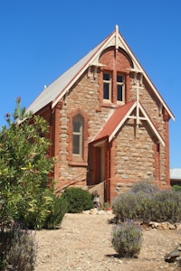 A quaint stone church with red brick accents on the windows and roof edges stands under a bright blue sky. The building has a steeply pitched roof with a small entrance door and cross-shaped wooden trim above it. Surrounding the church are various green and purple shrubs and dry, rocky ground.