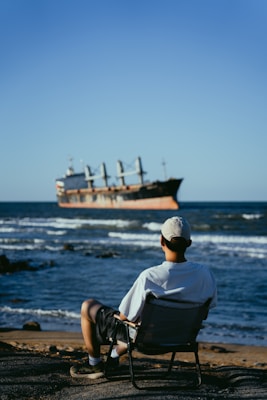 A person in a white shirt and cap sits on a beach chair facing the ocean, watching a large cargo ship pass by in the distance. The scene exudes a sense of relaxation and contemplation, with calm waves and a clear blue sky.