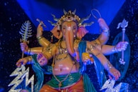 An artistic sculpture of a multi-armed deity with an elephant head, adorned with ornate jewelry, a crown, and holding various symbolic objects in each hand. The deity is draped in colorful garments, set against a starry blue background.