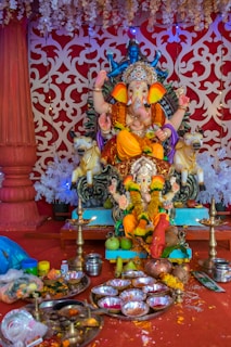 A colorful and ornate statue of the Hindu deity Ganesha is seated on an intricately designed throne. The statue is adorned with vibrant flowers, jewelry, and a crown. In front of the statue, there are offerings including fruits, a variety of items in metal plates, and traditional lamps. The background features an elaborate red and white patterned backdrop with hanging decorations.