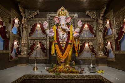 A richly decorated room features a large statue of the Hindu deity Ganesha, adorned with vibrant garlands and intricate jewelry. The backdrop consists of ornate carvings and luxurious drapes in deep red hues. Fruits and offerings are placed in front of the statue on the floor.