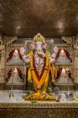 A detailed and ornate statue of the Hindu deity Ganesha, adorned with vibrant garlands of flowers. The statue is placed in an elaborately decorated shrine with intricate carvings and rich, warm colors. Offerings of fruits and incense are placed in front of the statue on a marble platform.