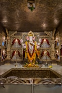 A beautifully adorned statue of a deity, decorated with vibrant garlands of marigolds and other flowers. The statue is placed in a sanctum with ornate carvings and red velvet drapes, surrounded by offerings of fruits and flowers. The setting has a richly decorated backdrop with intricate gold designs and traditional lamps.