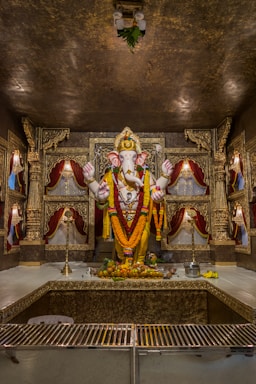 A beautifully adorned statue of a deity, decorated with vibrant garlands of marigolds and other flowers. The statue is placed in a sanctum with ornate carvings and red velvet drapes, surrounded by offerings of fruits and flowers. The setting has a richly decorated backdrop with intricate gold designs and traditional lamps.