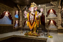 A large, intricately decorated statue of the Hindu deity Ganesha is adorned with vibrant flower garlands and set within an ornate room. Surrounding the statue are walls with detailed gold carvings and fabric drapes in rich red tones. Offerings, including fruits and a metal pot, are placed at the base of the statue.