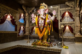 A large, intricately decorated statue of the Hindu deity Ganesha is adorned with vibrant flower garlands and set within an ornate room. Surrounding the statue are walls with detailed gold carvings and fabric drapes in rich red tones. Offerings, including fruits and a metal pot, are placed at the base of the statue.