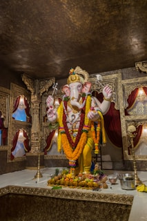 A detailed idol of Lord Ganesha adorned with colorful garlands and ornaments stands on a decorated platform. The background features intricately designed golden and maroon drapes and panels. Offerings like fruits and flowers are placed at the base of the idol, and there are traditional lamps and metalware around it.