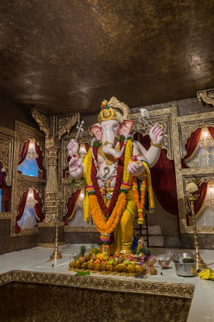 A detailed idol of Lord Ganesha adorned with colorful garlands and ornaments stands on a decorated platform. The background features intricately designed golden and maroon drapes and panels. Offerings like fruits and flowers are placed at the base of the idol, and there are traditional lamps and metalware around it.