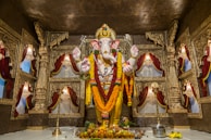 A grand and ornate statue of a deity with an elephant head, adorned with garlands and decorations, stands at the center of a lavish temple setting. The background features richly crafted gold-colored carvings and draped red curtains, creating a sacred and majestic atmosphere. Offerings such as fruits and a metal pot are placed at the base of the statue.