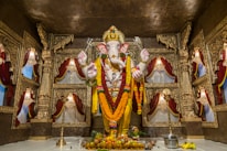 A grand and ornate statue of a deity with an elephant head, adorned with garlands and decorations, stands at the center of a lavish temple setting. The background features richly crafted gold-colored carvings and draped red curtains, creating a sacred and majestic atmosphere. Offerings such as fruits and a metal pot are placed at the base of the statue.