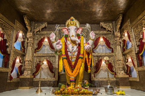 A grand and ornate statue of a deity with an elephant head, adorned with garlands and decorations, stands at the center of a lavish temple setting. The background features richly crafted gold-colored carvings and draped red curtains, creating a sacred and majestic atmosphere. Offerings such as fruits and a metal pot are placed at the base of the statue.