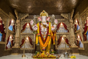 A grand and ornate statue of a deity with an elephant head, adorned with garlands and decorations, stands at the center of a lavish temple setting. The background features richly crafted gold-colored carvings and draped red curtains, creating a sacred and majestic atmosphere. Offerings such as fruits and a metal pot are placed at the base of the statue.