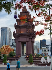 A monument with intricate architectural designs stands prominently, surrounded by flags and fountains. In the background are modern high-rise buildings. Red flowers frame the scene from above. Two people walk in the foreground.