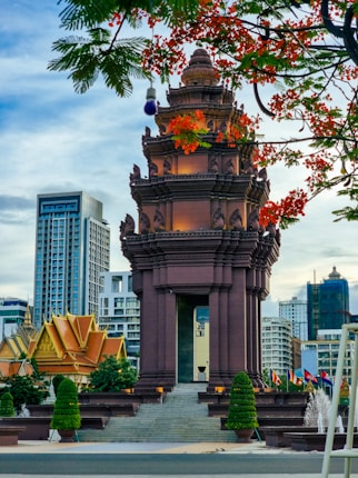 A historical monument with intricate architectural features stands prominently against a backdrop of modern high-rise buildings. The foreground features lush foliage and vibrant red flowers, adding a splash of color. Flags are displayed alongside carefully manicured trees, creating a blend of nature and urban elements.