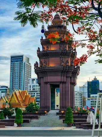 A historical monument with intricate architectural features stands prominently against a backdrop of modern high-rise buildings. The foreground features lush foliage and vibrant red flowers, adding a splash of color. Flags are displayed alongside carefully manicured trees, creating a blend of nature and urban elements.