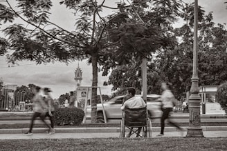 A man sitting in a wheelchair on a sidewalk observes the blurred motion of people walking briskly past him. The setting is an urban park with trees and lampposts, and a road with moving cars can be seen in the background.