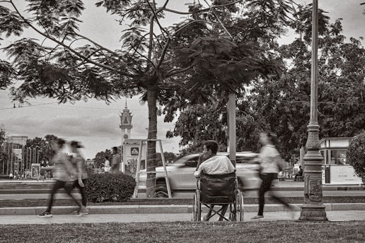 A man sitting in a wheelchair on a sidewalk observes the blurred motion of people walking briskly past him. The setting is an urban park with trees and lampposts, and a road with moving cars can be seen in the background.
