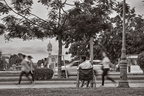 A man sitting in a wheelchair on a sidewalk observes the blurred motion of people walking briskly past him. The setting is an urban park with trees and lampposts, and a road with moving cars can be seen in the background.