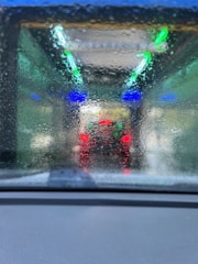 A blurred and wet windshield with streaks of water, through which colorful lights can be seen. There are vivid blue and green lights overhead, and red tail lights of a vehicle in front are visible, indicating the setting is a car wash tunnel.