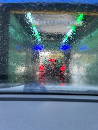 A blurred and wet windshield with streaks of water, through which colorful lights can be seen. There are vivid blue and green lights overhead, and red tail lights of a vehicle in front are visible, indicating the setting is a car wash tunnel.