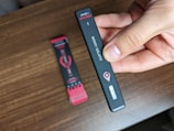 A hand holding a black vape pen labeled 'Berry Gelato' with a small window indicator. In the background, there is a partially visible packaging box on a wooden surface.