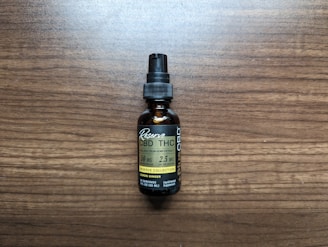 A small brown bottle with a black dropper cap labeled as a full spectrum hemp extract, specifically CBD and THC tincture. The label includes the dosage information as 50 mg per serving with 2.5 mg of THC per serving, and it mentions lemon ginger flavor. The bottle is placed on a wooden surface.