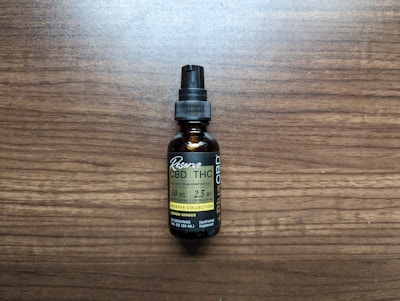 A small brown bottle with a black dropper cap labeled as a full spectrum hemp extract, specifically CBD and THC tincture. The label includes the dosage information as 50 mg per serving with 2.5 mg of THC per serving, and it mentions lemon ginger flavor. The bottle is placed on a wooden surface.