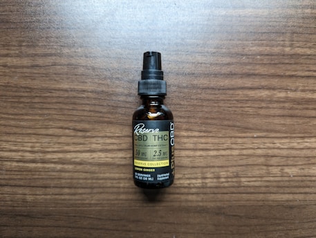 A small brown bottle with a black dropper cap labeled as a full spectrum hemp extract, specifically CBD and THC tincture. The label includes the dosage information as 50 mg per serving with 2.5 mg of THC per serving, and it mentions lemon ginger flavor. The bottle is placed on a wooden surface.