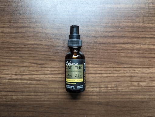 A small brown bottle with a black dropper cap labeled as a full spectrum hemp extract, specifically CBD and THC tincture. The label includes the dosage information as 50 mg per serving with 2.5 mg of THC per serving, and it mentions lemon ginger flavor. The bottle is placed on a wooden surface.