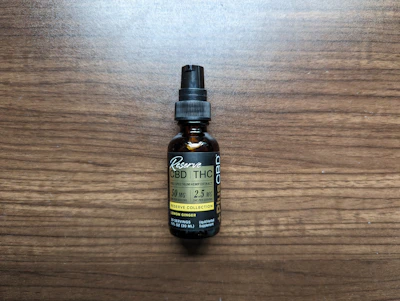 A small brown bottle with a black dropper cap labeled as a full spectrum hemp extract, specifically CBD and THC tincture. The label includes the dosage information as 50 mg per serving with 2.5 mg of THC per serving, and it mentions lemon ginger flavor. The bottle is placed on a wooden surface.