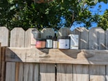 Several containers of CBD products are displayed on a wooden fence. The collection includes gummy jars, oil bottles, and boxes with labels. The background features trees and a clear blue sky.