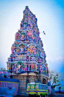A vibrant and intricately decorated Hindu temple tower adorned with colorful sculptures of deities, mythological figures, and ornate patterns. The structure is set against a clear blue sky, with a bus visible at the base.