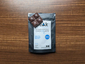 A package of strain-specific chocolate labeled as containing 86 mg of cannabinoids, placed on a wooden surface. A small dark chocolate piece rests partially on the package, which features 'Special Sauce' in blue text. The packaging indicates it is 70% dark hemp chocolate.