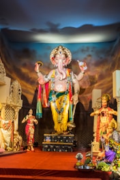 A large and intricate statue of Lord Ganesha is prominently displayed, adorned with vibrant garlands and colorful attire. The surroundings include additional smaller statues and offerings laid out on the floor. The backdrop features a dramatic, cloud-filled sky.