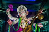 A colorful statue of the Hindu deity Ganesha adorned with intricate jewelry and garments. The figure holds a musical instrument and a pink flower, surrounded by decorative elements and vibrant drapery.