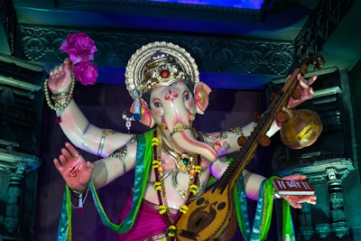 A colorful statue of the Hindu deity Ganesha adorned with intricate jewelry and garments. The figure holds a musical instrument and a pink flower, surrounded by decorative elements and vibrant drapery.