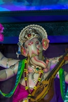 A detailed statue of a deity with an elephant head adorned with an ornate golden crown and intricate jewelry. The statue is elaborately decorated in vibrant colors, including pink, green, and blue garments. The deity holds a stringed musical instrument resembling a veena, symbolizing cultural and spiritual significance. The background features a swirling design with blue and purple hues.