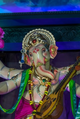 A detailed statue of a deity with an elephant head adorned with an ornate golden crown and intricate jewelry. The statue is elaborately decorated in vibrant colors, including pink, green, and blue garments. The deity holds a stringed musical instrument resembling a veena, symbolizing cultural and spiritual significance. The background features a swirling design with blue and purple hues.